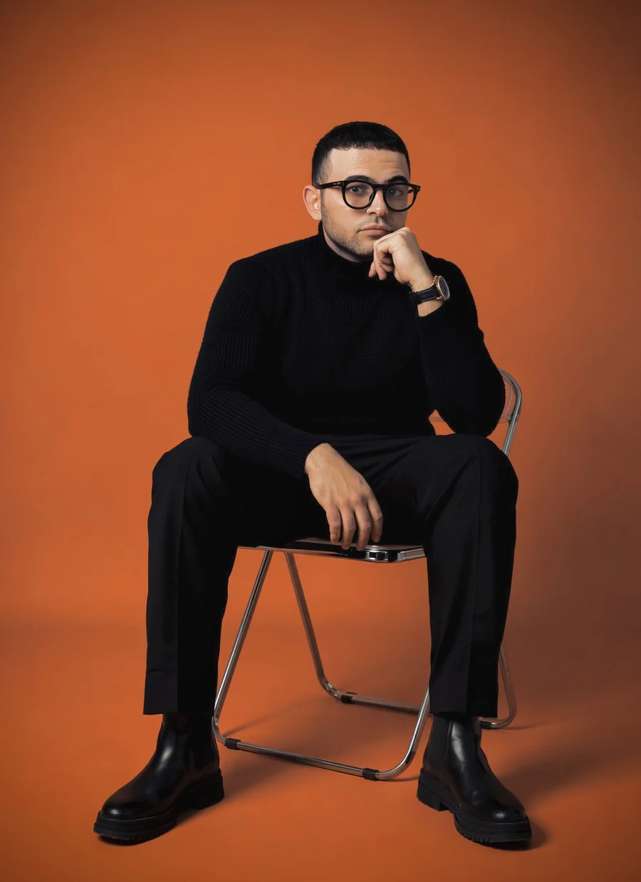 Ahmed Naeem in a black turtleneck, sitting on a folding chair against an orange backdrop